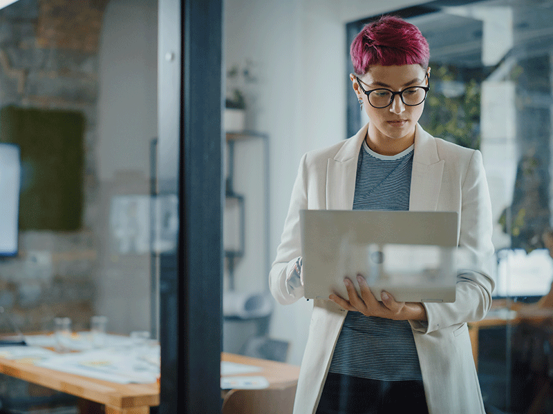 Modern Office: Portrait of Beautiful Authentic Specialist with Short Pink Hair Standing, Holding Laptop Computer, Concentrated on Her Work. Working on Design, Data Analysis, Plan Strategy