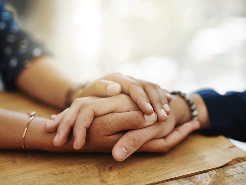 Holding hands, support and closeup with trust, solidarity and community on a home table. Therapy, diversity and gratitude of friends together with hope, respect and love for grief empathy and forgive