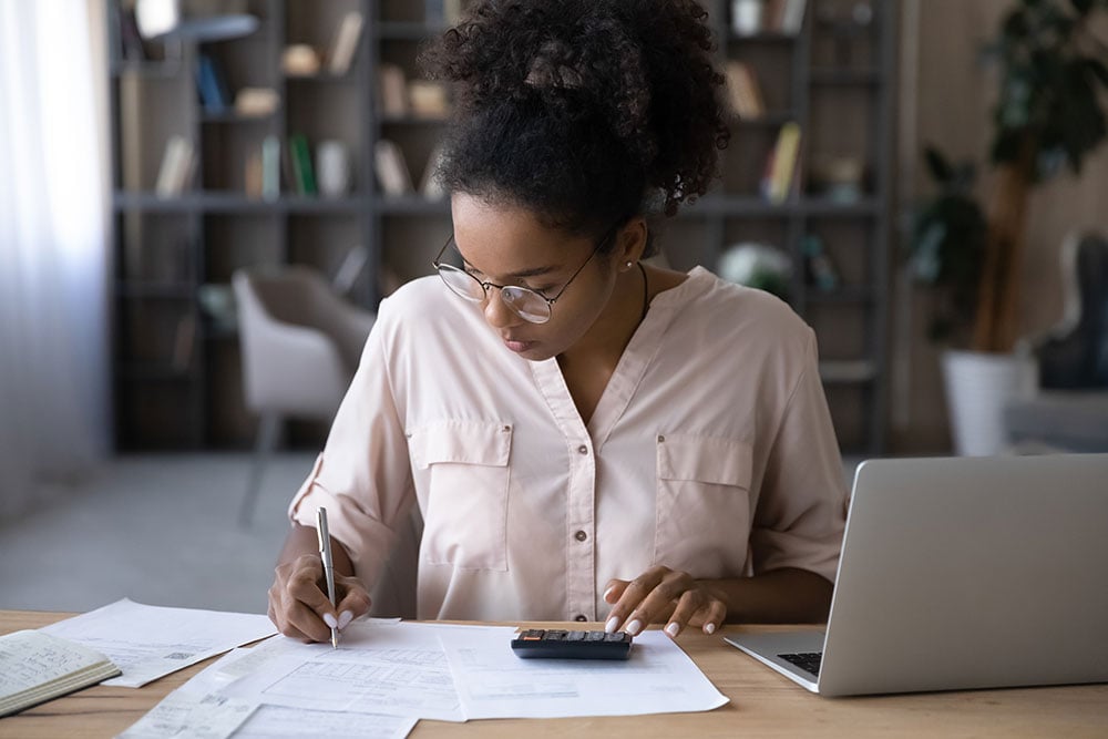 Serious young biracial woman sit at desk manage budget calculate on machine pay bills taxes online on laptop. Focused African American female count expenses expenditures on calculator. Save concept.