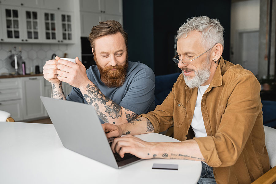 Tattooed gay couple with coffee using laptop near credit card at home.