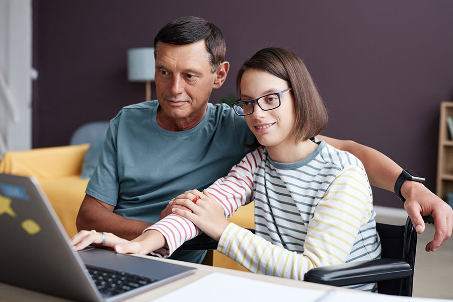 smiling teenage girl with disability using laptop computer at home with father helping